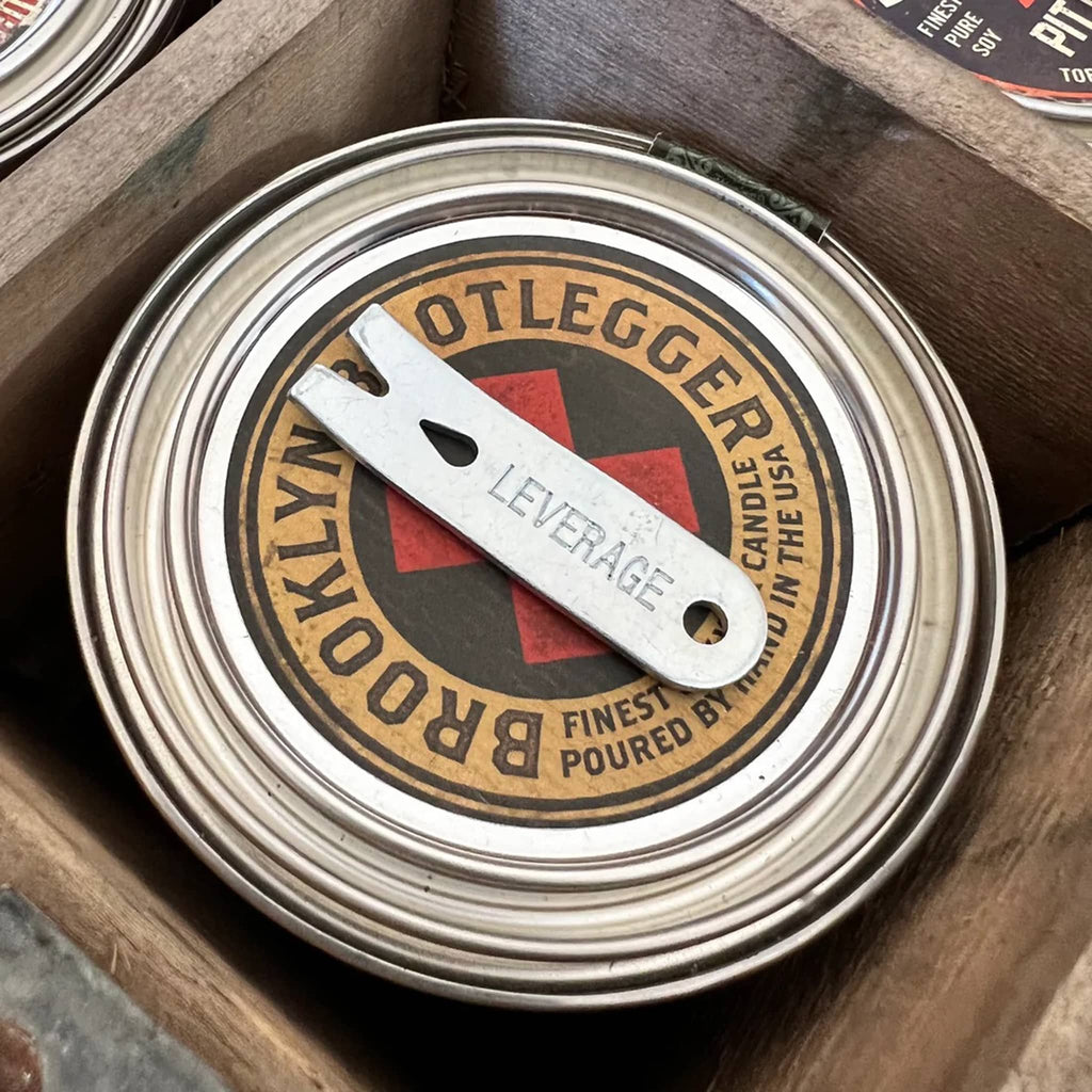 Top view of Buster Mandel Co. Brooklyn Bootlegger scented soy wax candle in metal tin with lid, candle name is on lid and a mini metal leverage crowbar to open the tin lid.