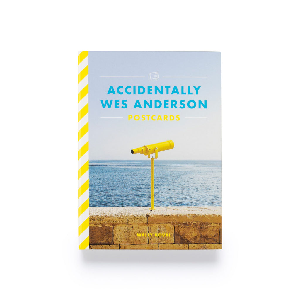 Cover of Accidentally Wes Anderson postcard book with a photo of a yellow telescope on a beige sea wall with a blue ocean and sky in background. The spine is yellow and white striped.