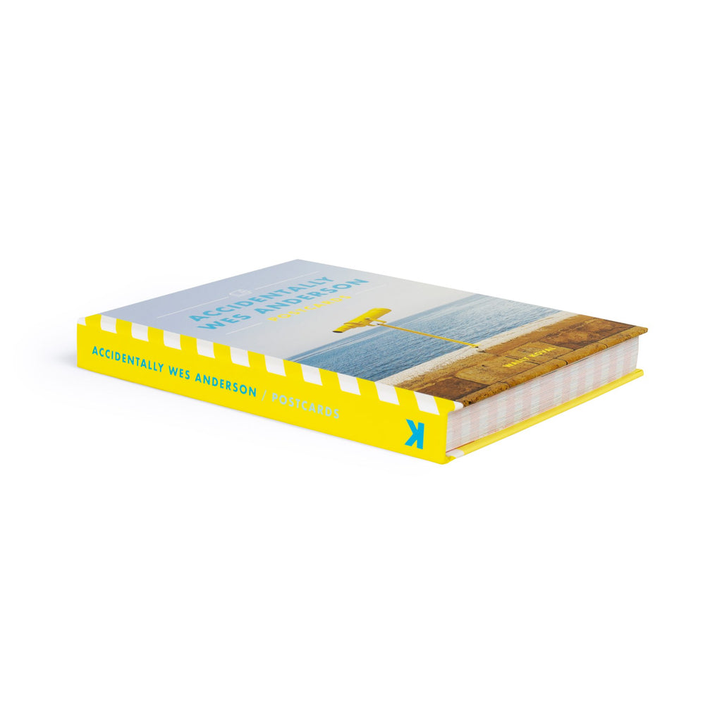 Accidentally Wes Anderson postcard book with a photo of a yellow telescope on a beige sea wall with a blue ocean and sky in background on the cover. The book is lying down on a white surface and the view shows the spine, front cover and bottom of the book. The spine is yellow with the book name in aqua blue.