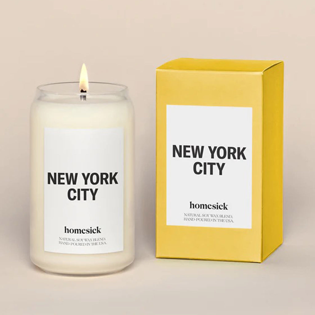 candle in clear glass can-shaped tumbler lit, white panel and "new york city" with "homesick" in black lettering beside yellow gift box with white panel and "new york city" and "homesick" with black lettering