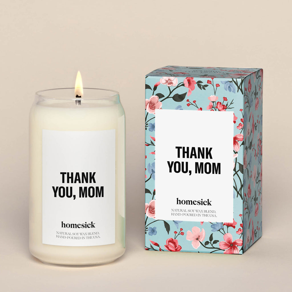 homesick soy wax candle in a can-shaped clear glass jar with white label and "thank you, mom" in black letters beside gift box packaging with pink and blue flowers on a blue background and "thank you, mom" in black letters on white box on front 
