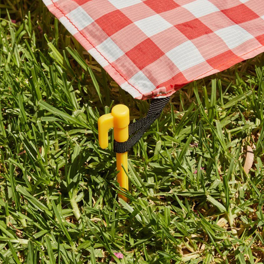 Kikkerland red & white gingham print ripstop nylon picnic blanket, close-up of corner with yellow stake in grass.