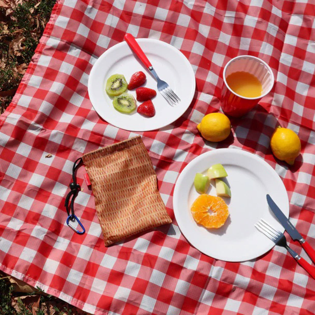 Kikkerland red & white gingham print ripstop nylon picnic blanket spread out on grass with plates of fruit and the  in included wicker print pouch.