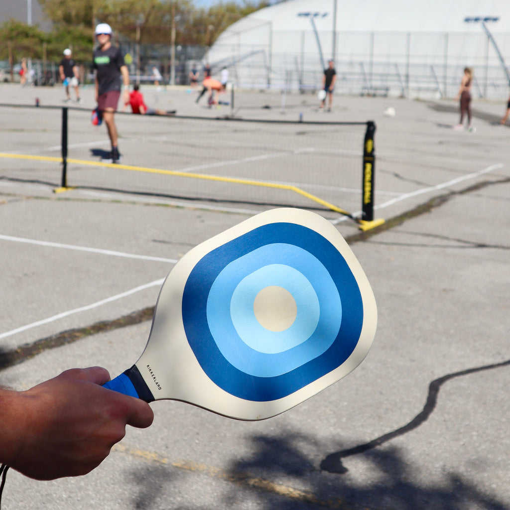 Kikkerland Pickleball Set in use on a court, a hand is holding a wood paddle with blue grip and ombre blue pattern.