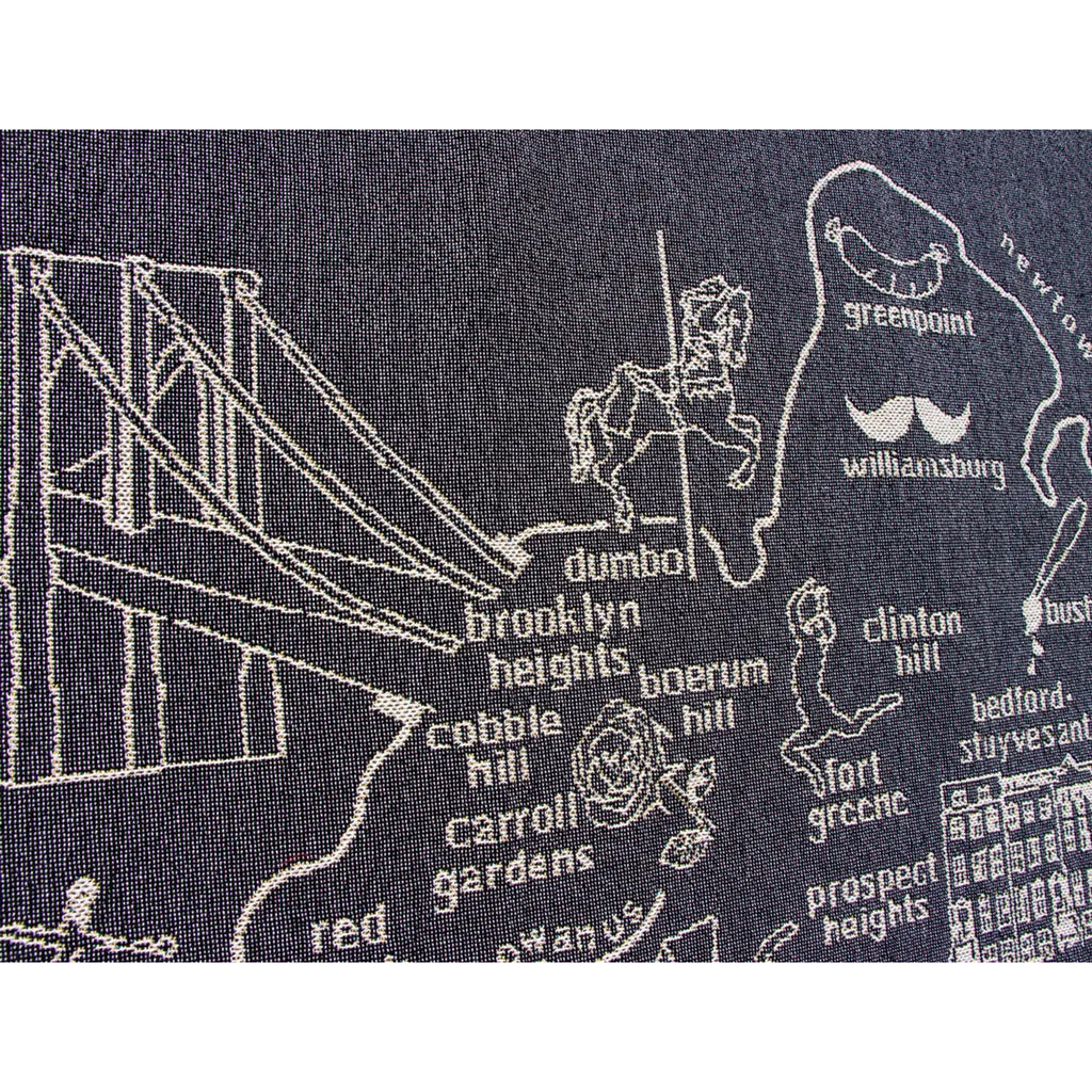 Detail of Dumbo, Cobble Hill, Boerum Hill and other neighborhood names on a Brooklyn-themed knit throw blanket, illustrations and wording in white on a navy blue backdrop.