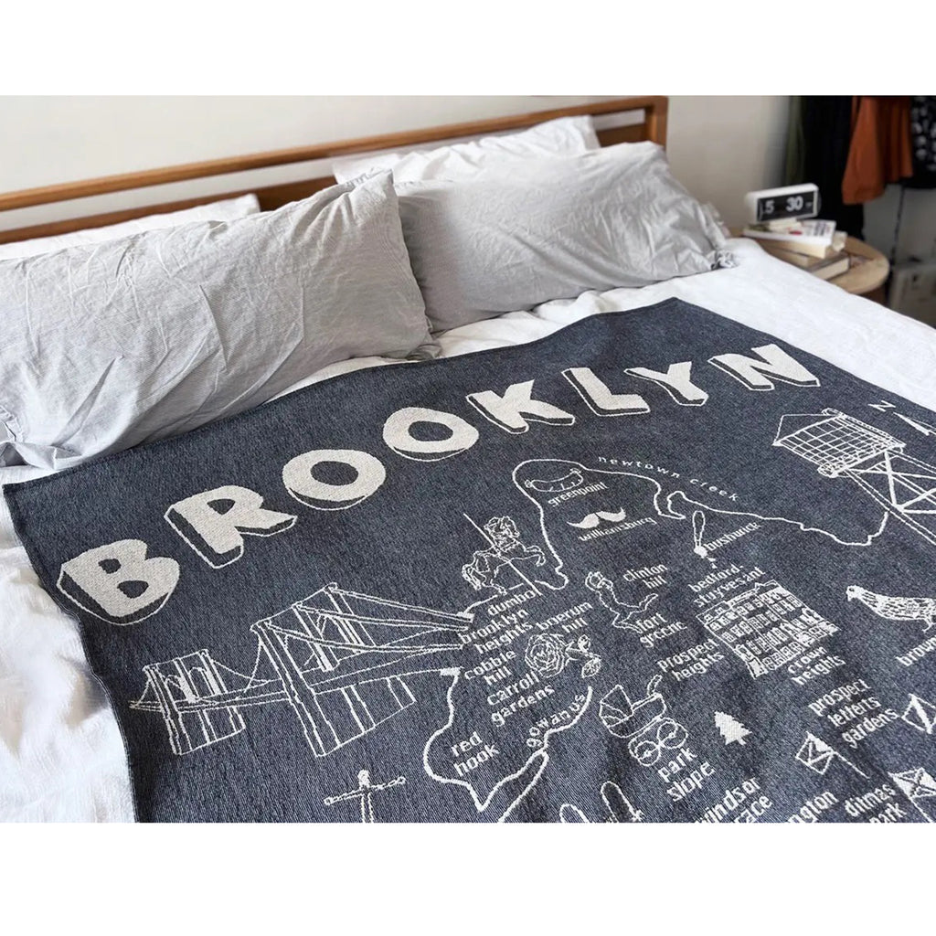 Detail of Brooklyn-themed knit throw blanket with illustrations and wording in white on a navy backdrop, blanket is spread on a bed with gray sheets.