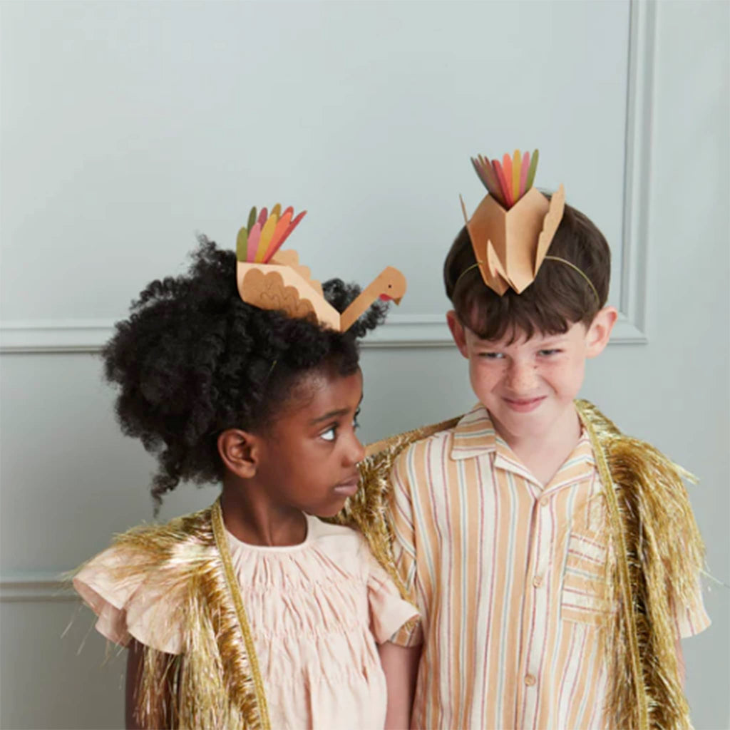 Folded paper party hat that looks like a turkey with gold foil beak and colorful tail feathers with a gold elastic headband on the heads of a boy and a girl.