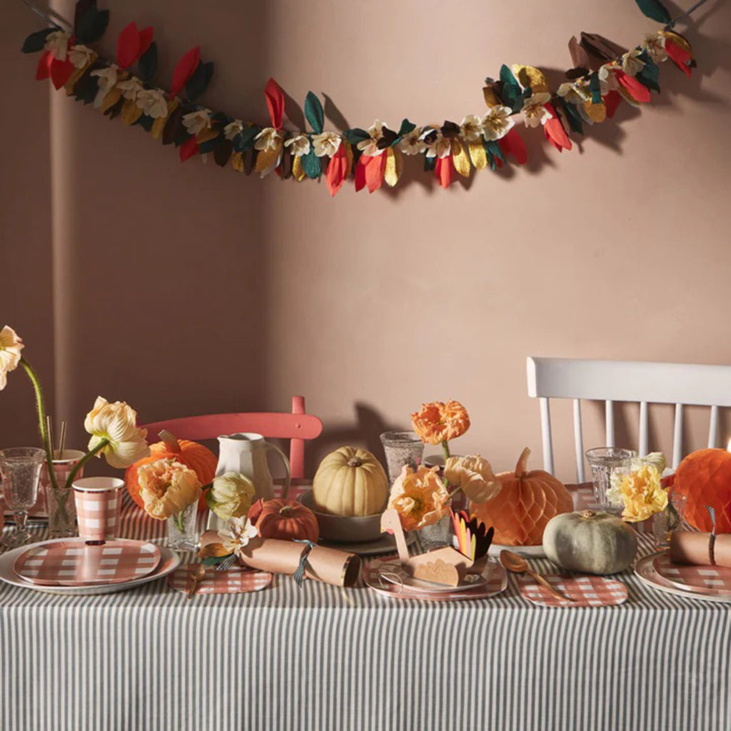 Crepe paper leaf and flower garland in fall colors of green, brown, orange and gold strung on a green gingham ribbon shown in a fall party setting with set table.