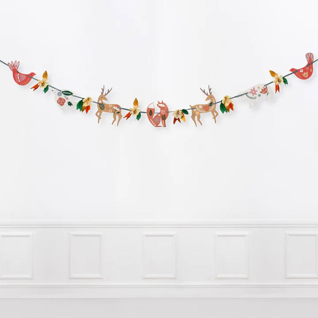 Woodland animals with folk art type illustrations and crepe paper flower and leaf clusters strung on a green gingham ribbon, shows full length against a white wall. 