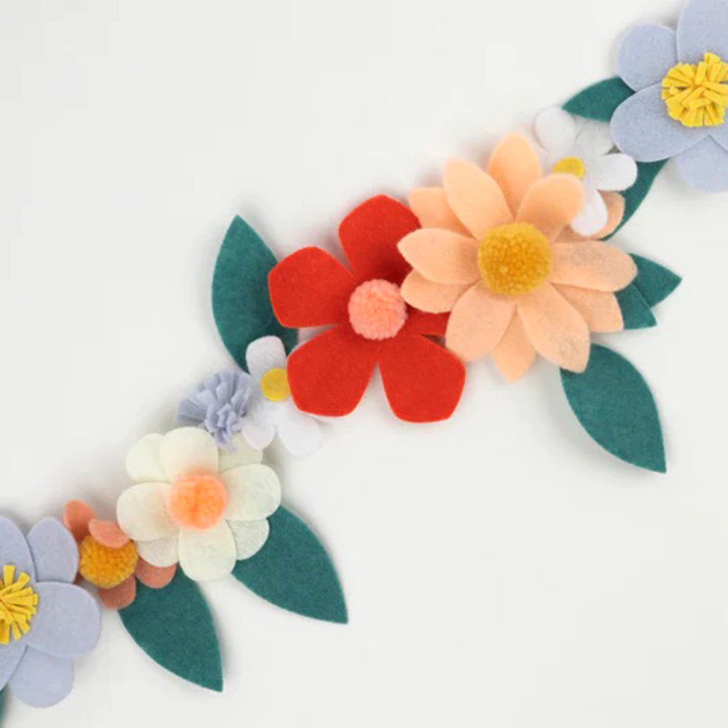 Meri Meri Felt Flower Garland with cream, light yellow, peach, coral, pink and pale blue flowers with pompom centers and green leaves, close-up detail view.
