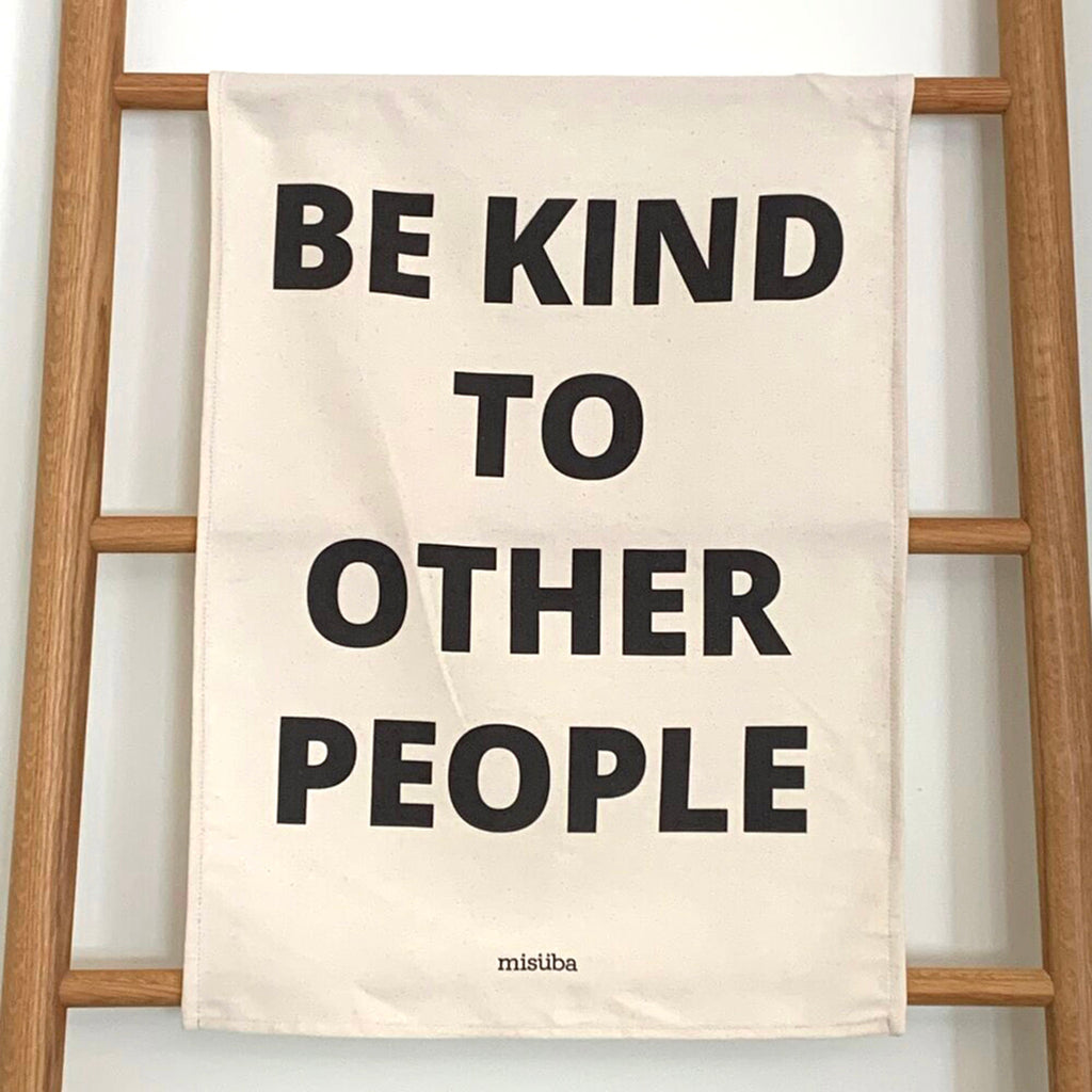 misuba unbleached cotton kitchen dish tea towel with "be kind to other people" in black letters handing on wood ladder