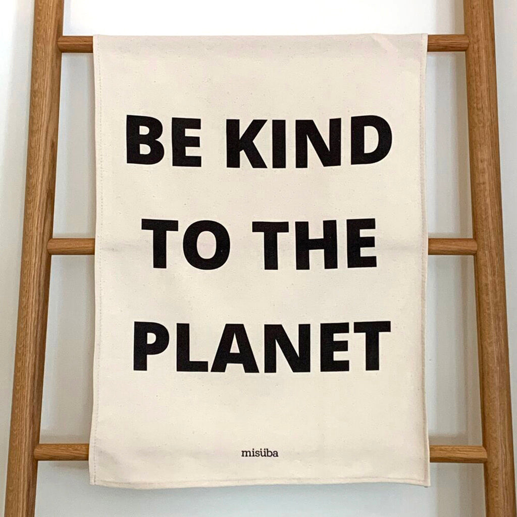 misuba unbleached cotton kitchen dish tea towel with "be kind to the planet" in black letters handing on wood ladder