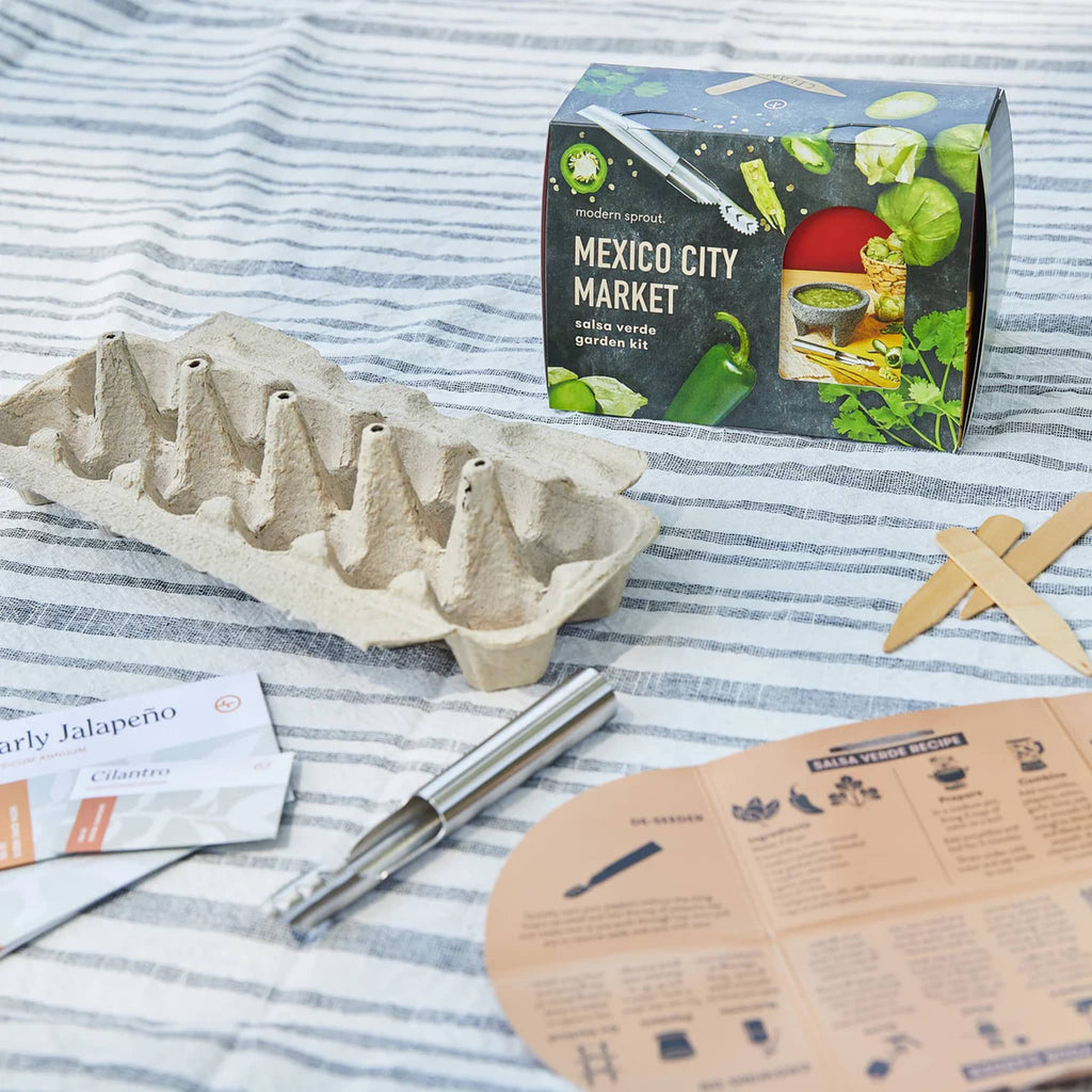 Front of packaging for the Mexico City Market Salsa Verde Garden Kit with seedling tray, pepper deseeder tool, wooden garden markers, seed packages and instructions on gray and white striped fabric.