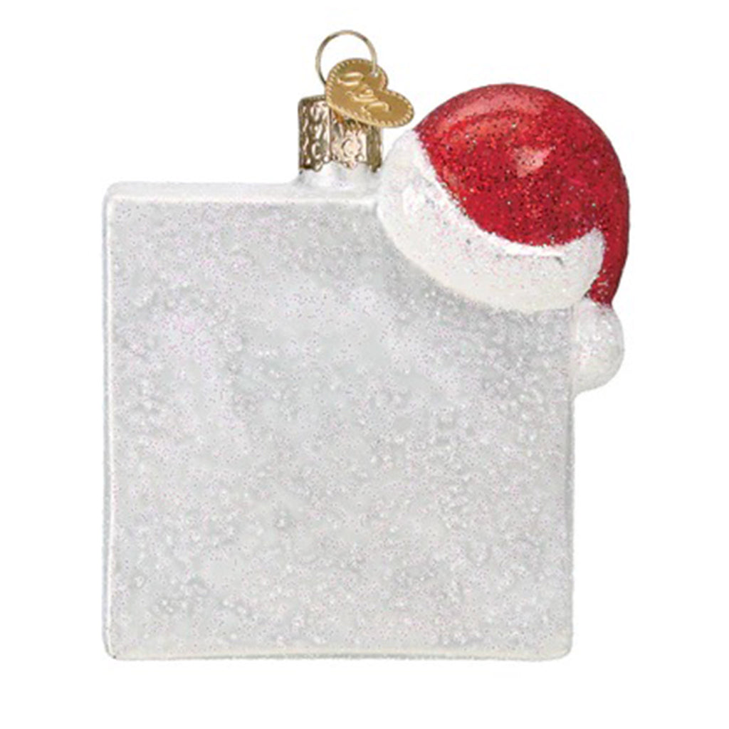 Back view of a glass holiday tree ornament with a white glitter square  and a santa hat hanging on the top right corner.