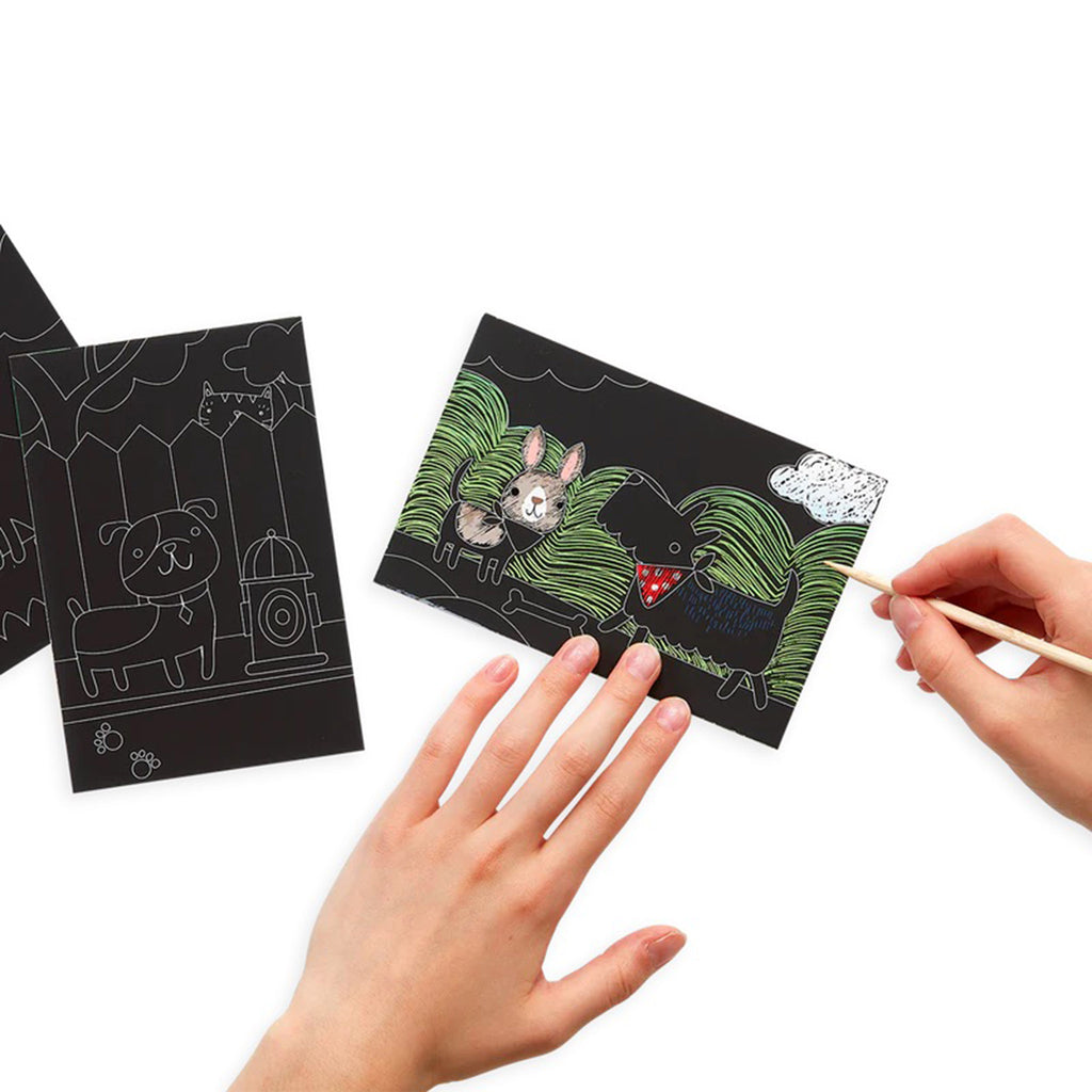 scratch & scribble mini kit playful pups guided artwork with left hand holding down black sheet of paper with a sketch of a chihuahua with a scottish terrier while the right hand is holding a wood scratching tool revealing some of the colors beneath