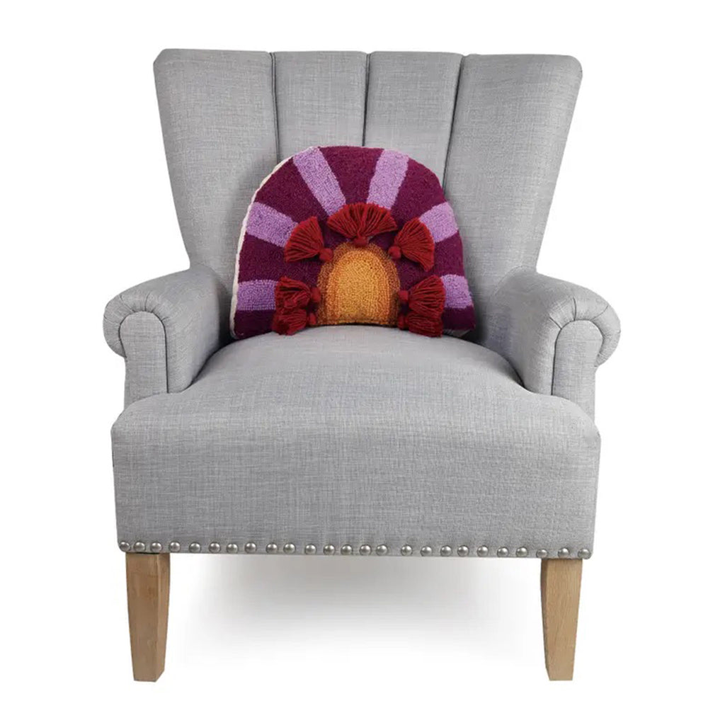 Peking Handicraft In Bloom arc shaped hooked wool throw pillow with burgundy and purple rays coming out from orange and yellow arches with maroon tassels on a gray tufted chair.