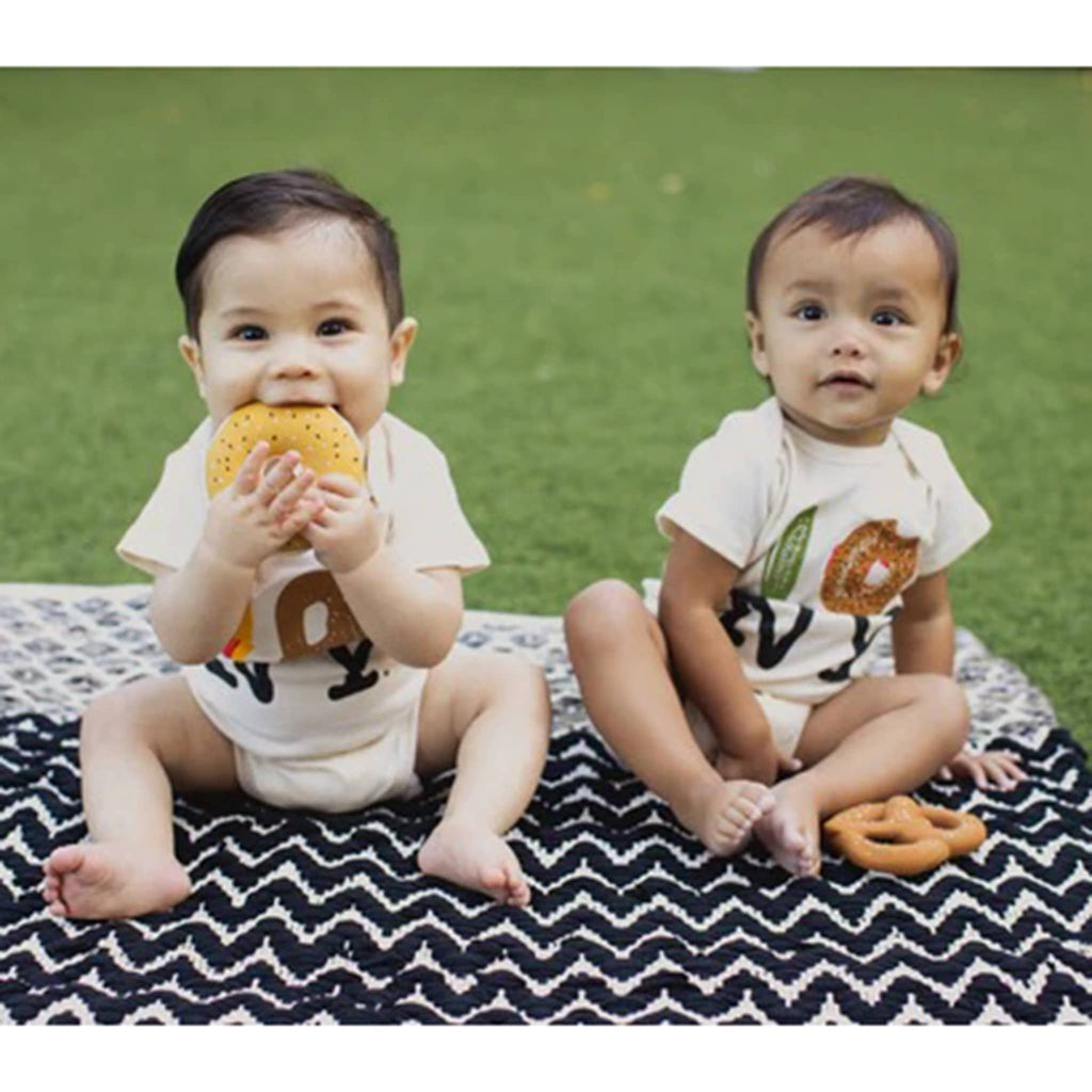 Soft pretzel and bagel with lox shaped natural rubber baby teether bath toys with babies on a black and white blanket with grass in the background.