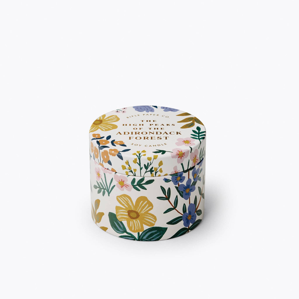 Scented candle in a metal tin with lid that features a muted floral print in yellow, pink, green and blue on a white background. Lid is on.