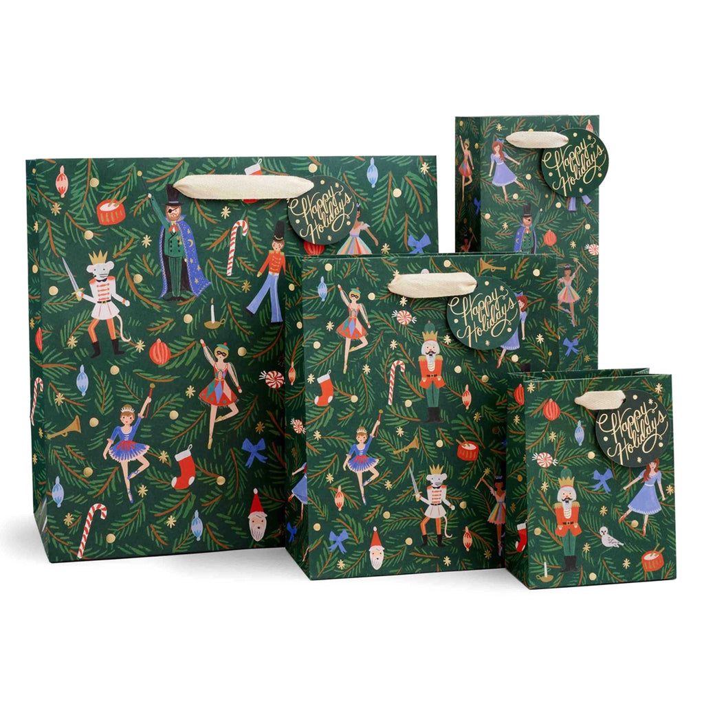 4 sizes of gift bags with characters from the Nutcracker Ballet on them on a backdrop of dark green with evergreen boughs, satin ribbon handles and a round gift tag.