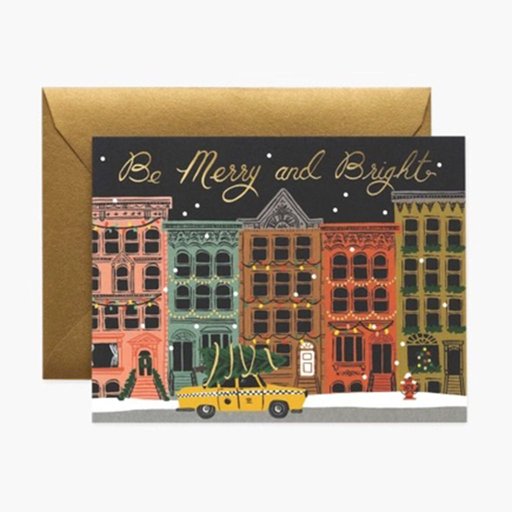 Front of holiday card with a row of colorful brownstones behind a taxi with a tree on top with "be merry and bright" in gold foil in the night sky with matching metallic gold envelope.