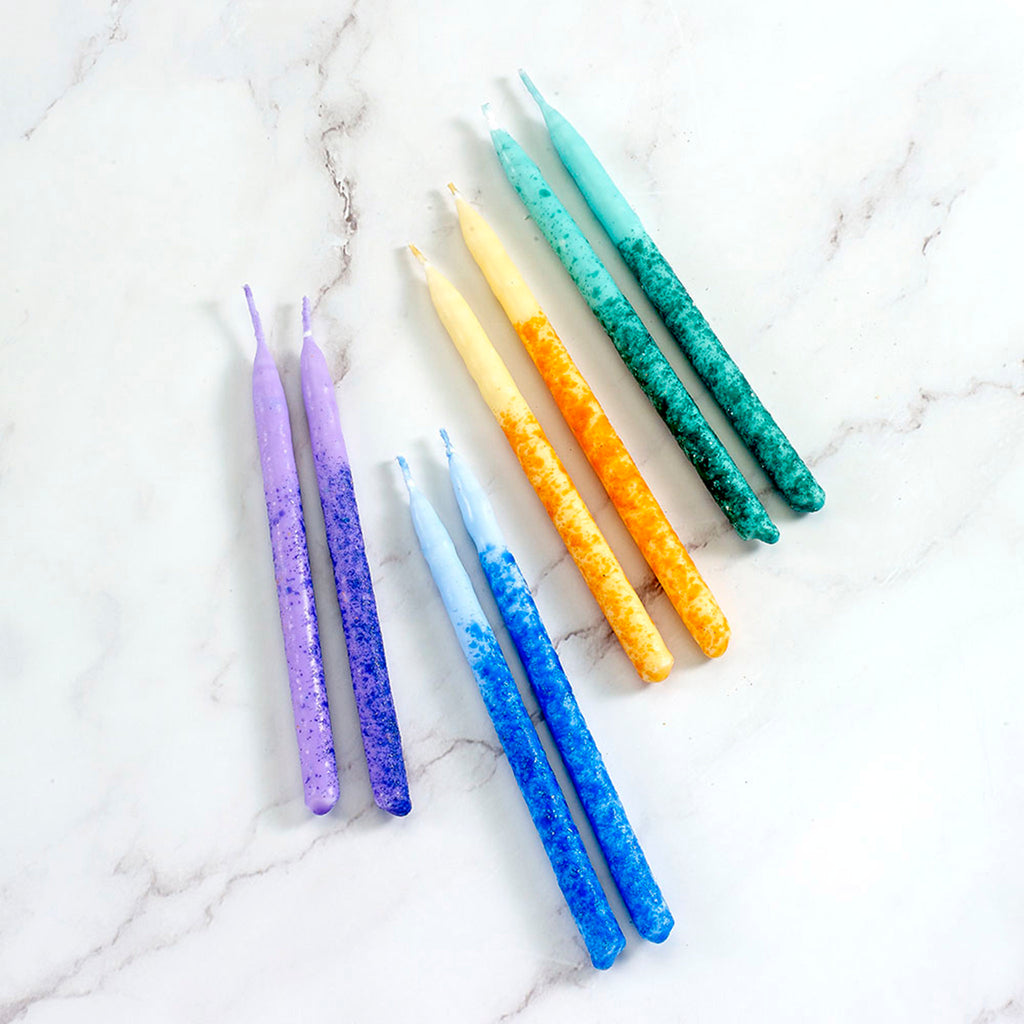 Premium multicolor chanukah hanukkah menorah candles in purple, blue, yellow/orange and green on a white marble surface.
