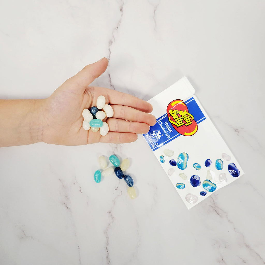 Jelly Belly assorted blue & white jelly beans in a white pouch with Happy Chanukah on the front and dreidel illustrations. Bag is open and a hand is holding a mix of jelly beans.