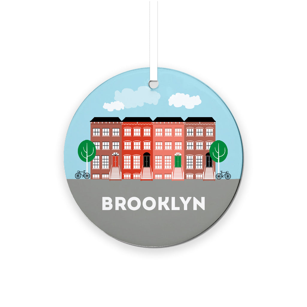 Round ceramic holiday ornament with a row of pink, red and brown brownstones against a light blue sky with white clouds and a gray foreground with "brooklyn" in white lettering.