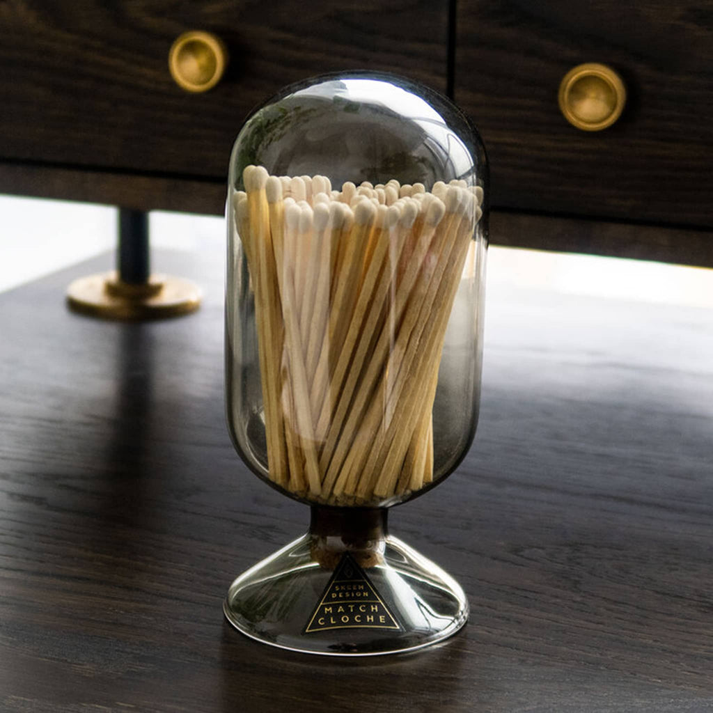 Vertical oblong oval shaped smoke gray glass vessel with circular base and filled with white tipped wood safety matches on dark wood desk.