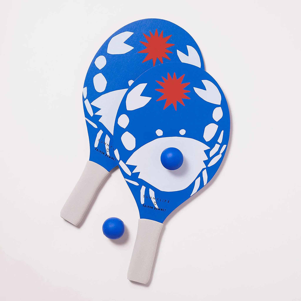 sunnylife 2 beach bats in whack print with white foam handles and blue paddles with a white crab and red sun along with 2 blue balls