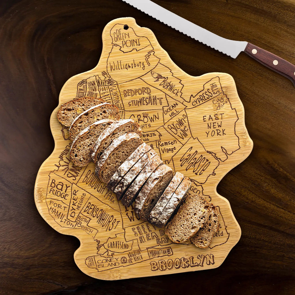 Brooklyn shaped bamboo serving board that is laser engraved with the neighborhood shapes and names and "brooklyn" at the bottom. Board has sliced brown bread on it and a wood handled serrated bread knife beside it on a dark wood background.