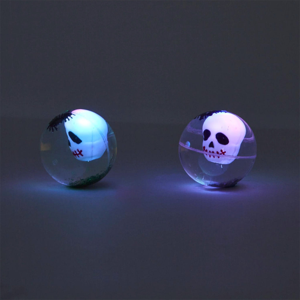 Front view of rubber bouncing balls with a light up skull, little black spiders and green or silver glitter on a black background in the dark.