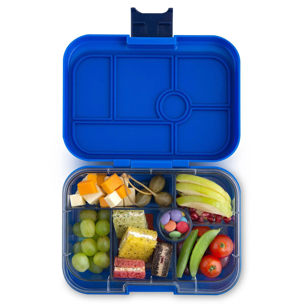 Yumbox lunch box in royal neptune blue, lid open showing clear inner tray divided into 6 compartments. Each compartment has food in it: white and orange cheese cubes, olives, sliced apples, pomegranate seeds, snap peas, cherry tomatoes, chocolate candies, green grapes and little bite-sized sandwiches with filling and different colored bread (white, yellow, brown and pink).