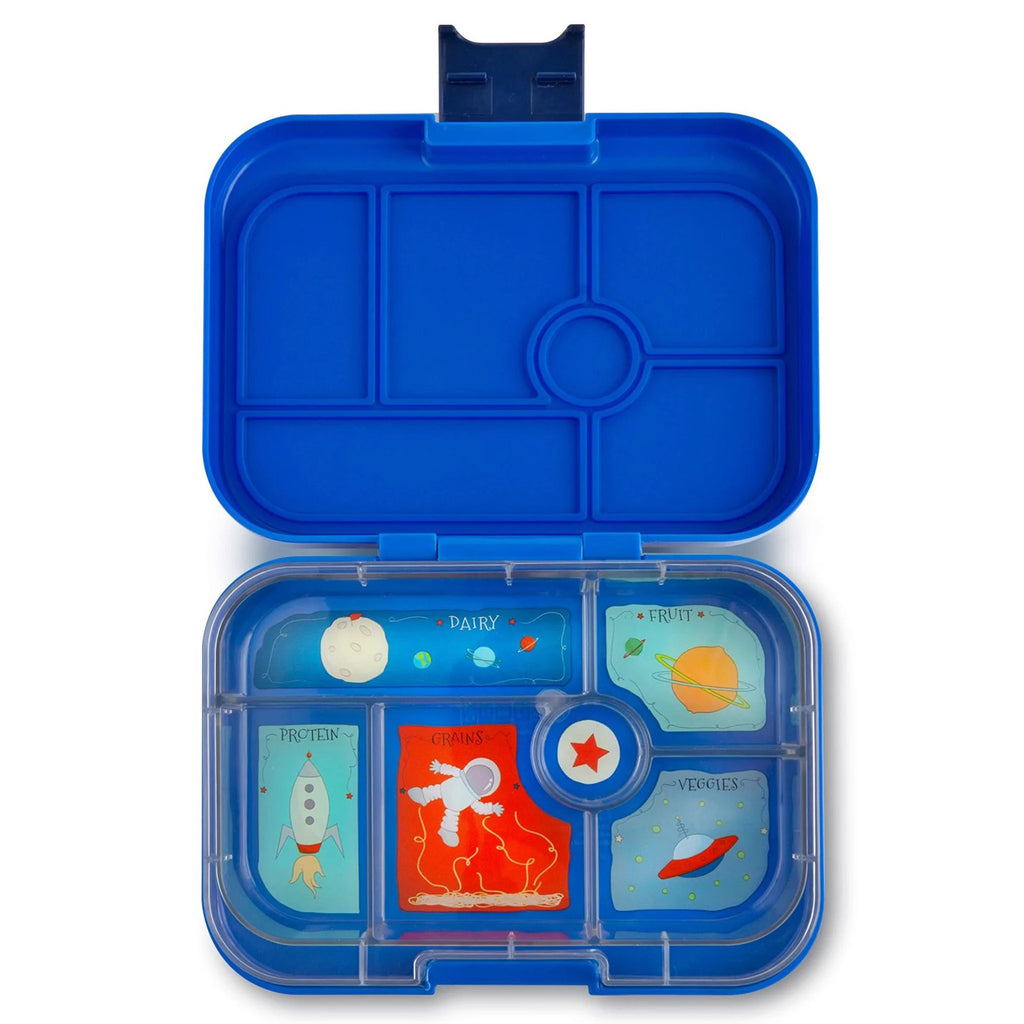Yumbox lunch box in royal neptune blue, lid open showing clear inner tray divided into 6 compartments. Each compartment has an illustration (planets, moon, earth, astronaut, spaceship, red star and rocket) and the 5 larger compartments each have a label of dairy, protein, grains, fruit or veggies in light blue or dark blue script letters.