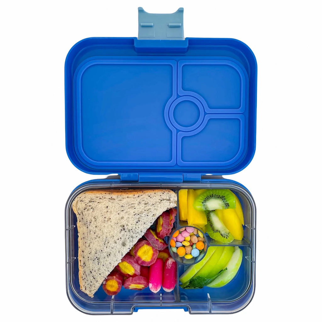 yumbox lunch box in royal blue, lid open showing clear inner tray divided into 4 compartments with a 1/2 sandwich, cut vegetables, chocolate candies, sliced apples, kiwi and mango